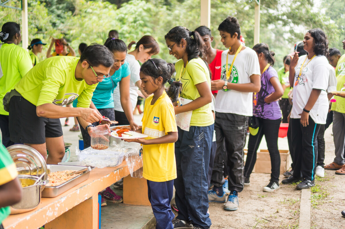 Care2Run volunteers serving a healthy community meal to youth participants during an inclusive CSR opportunity sports camp in Malaysia.