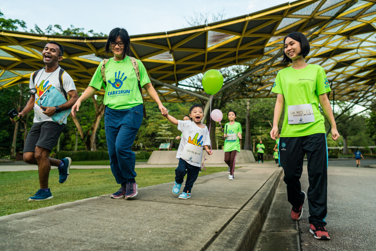 A young child holding hands with a Care2Run participants while walking with a green balloon during an inclusive sports CSR opportunity event in Malaysia.